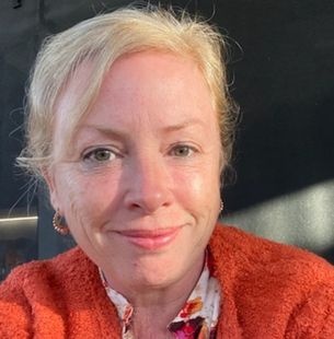 A headshot of a blonde woman wearing an orange jacket over a flowery shirt