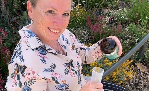 Katrina is sitting surrounded by colourful flowers, she is holding a dark bottle and a graduated cylinder